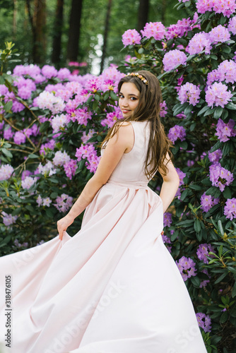 Beautiful young brunette with a smile and big brown eyes in blooming gardens of pink rhododendrons in a long pink delicate dress