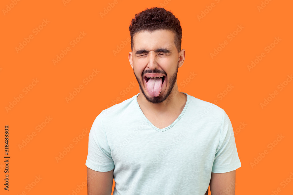 Portrait of childish carefree brunette man with beard in white t-shirt showing out tongue and closing eyes with naughty disobedient grimace, making face. studio shot isolated on orange background Portrait of childish carefree brunette man with beard in white t-shirt showing out tongue and closing eyes with naughty disobedient grimace, making face. studio shot isolated on orange background