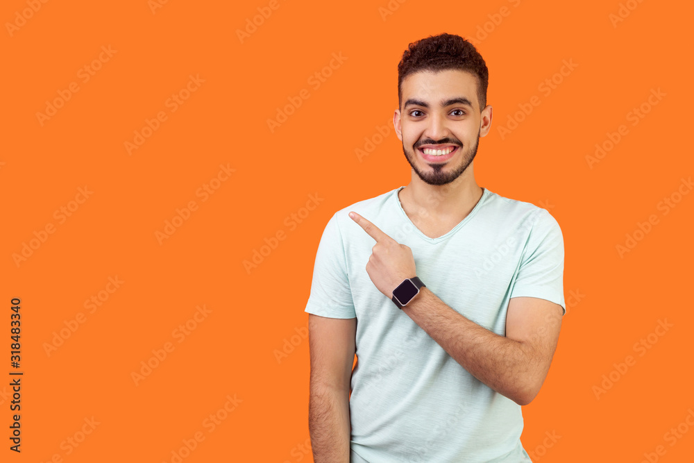 Look here! Portrait of positive friendly brunette man with beard in casual white t-shirt standing pointing at left, empty space for text, advertise. indoor studio shot isolated on orange background - obrazy, fototapety, plakaty 
