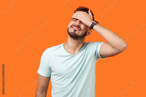 Facepalm. Portrait of desperate brunette man with beard in white t-shirt covering face with hand, feeling sorry and blaming himself for the mistake. indoor studio shot isolated on orange background photo