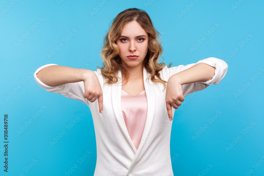 Fototapeta premium Here and right now! Portrait of strict serious woman boss with wavy hair in white jacket pointing down with angry face, giving immediate instructions, orders. studio shot isolated on blue background