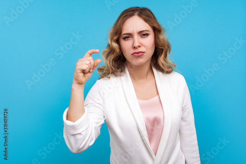 Some more please. Portrait of displeased woman with wavy hair in white jacket making a little bit gesture and frowning face, measuring small size. indoor studio shot isolated on blue background