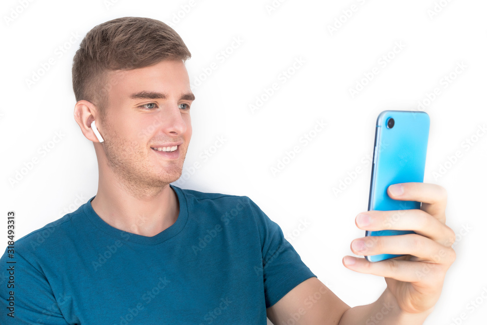 Handsome happy guy, young beautiful man smiling, looking at cell mobile phone in wireless earphones. Listening to music, talking, having conversation by video call, taking selfie, isolated on white. Handsome happy guy, young beautiful man smiling, looking at cell mobile phone in wireless earphones. Listening to music, talking, having conversation by video call, taking selfie, isolated on white.