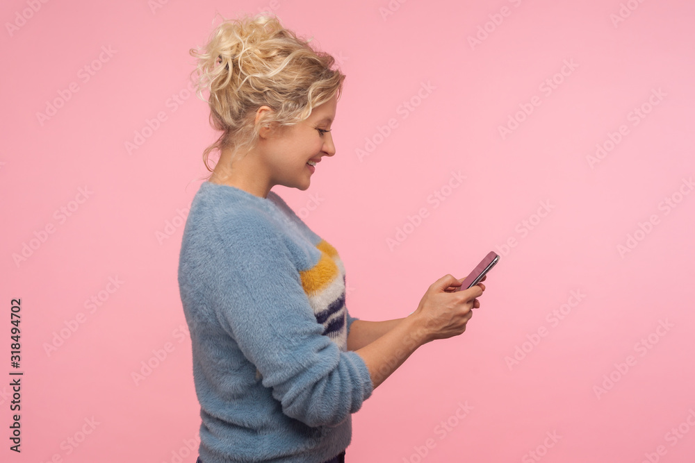 Side view of cheerful cute woman with curly hair in sweater chatting in cellphone, reading pleasant message and smiling charmingly, using mobile phone. indoor studio shot isolated on pink background Side view of cheerful cute woman with curly hair in sweater chatting in cellphone, reading pleasant message and smiling charmingly, using mobile phone. indoor studio shot isolated on pink background
