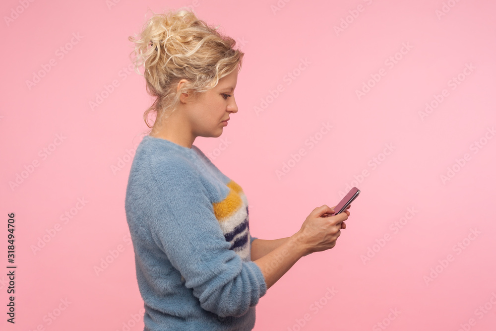Side view of serious pretty woman with short curly hair in warm sweater chatting in cellphone, reading message, using mobile phone with attentive look. indoor studio shot isolated on pink background Side view of serious pretty woman with short curly hair in warm sweater chatting in cellphone, reading message, using mobile phone with attentive look. indoor studio shot isolated on pink background