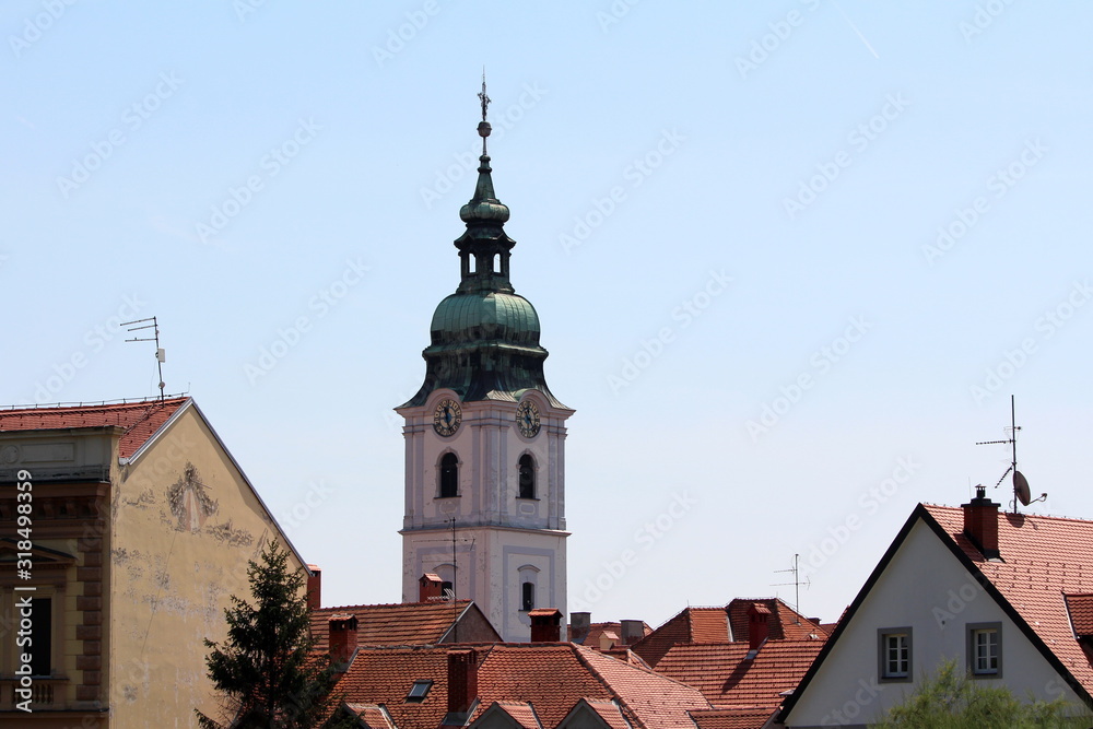 Church bell tower with analogue clocks on all sides and large metal cross on top covered with green metal tiles rising high above city family houses rooftops on clear blue sky background