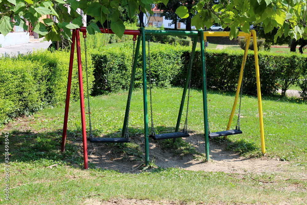 Fototapeta premium Colorful outdoor public playground equipment metal swing with three plastic seats in shade of tall old tree surrounded with grass and hedge in local public park