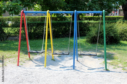 Front view of colorful outdoor public playground equipment swing with three plastic seats in shade of tall old trees surrounded with grass and hedge in local public park