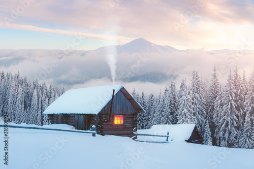 Fantastic winter landscape with wooden house in snowy mountains. Smoke comes from the chimney of snow covered hut. Christmas holiday and winter vacations concept