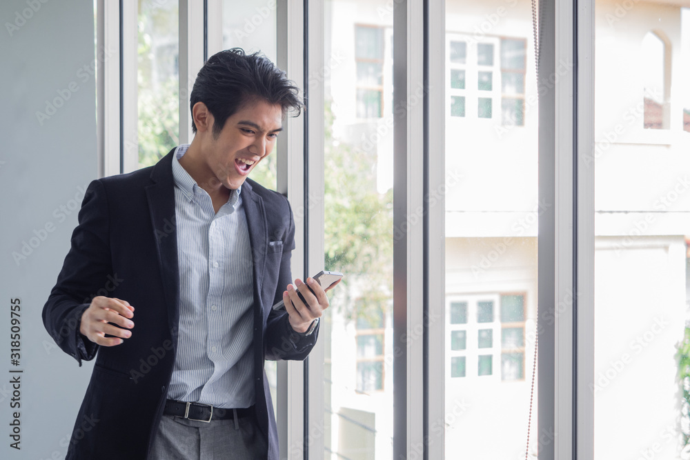 Handsome Asian businessman standing by the window uses the internet and social media on his smartphone to work in the office.He is happy because job is successful.Garden background with green trees. Handsome Asian businessman standing by the window uses the internet and social media on his smartphone to work in the office.He is happy because job is successful.Garden background with green trees.