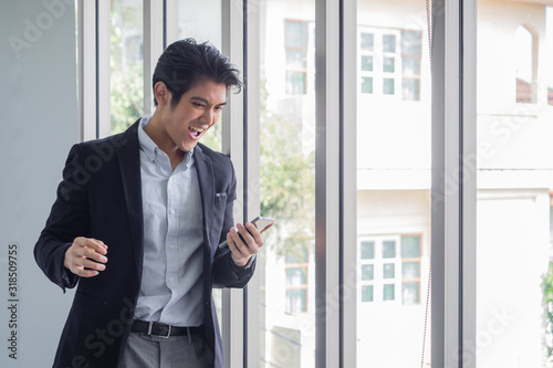 Handsome Asian businessman standing by the window uses the internet and social media on his smartphone to work in the office.He is happy because job is successful.Garden background with green trees.