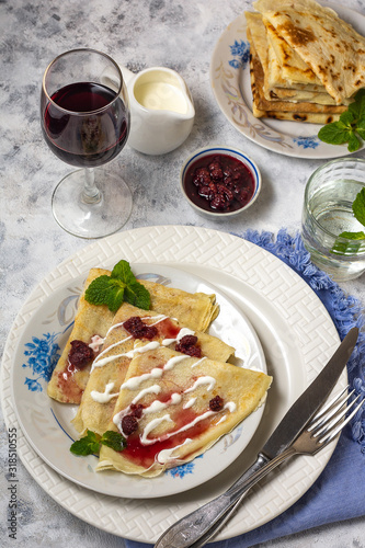 Pancakes on a plate with sour cream and raspberry jam and sprigs of mint with appliances on a gray background with a glass of wine and a glass of water vertical arrangement