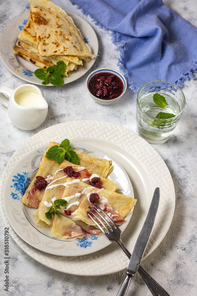 Pancakes on a plate with sour cream and raspberry jam and sprigs of mint with devices on a gray background with a glass of water vertical arrangement