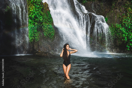 Young slim brunette woman with curly hair enjoying in lagoon of huge tropical waterfall Banyumala in Bali. Wearing in black swimsuit. Happy vacations in Indonesia. Wanderlust travel concept.