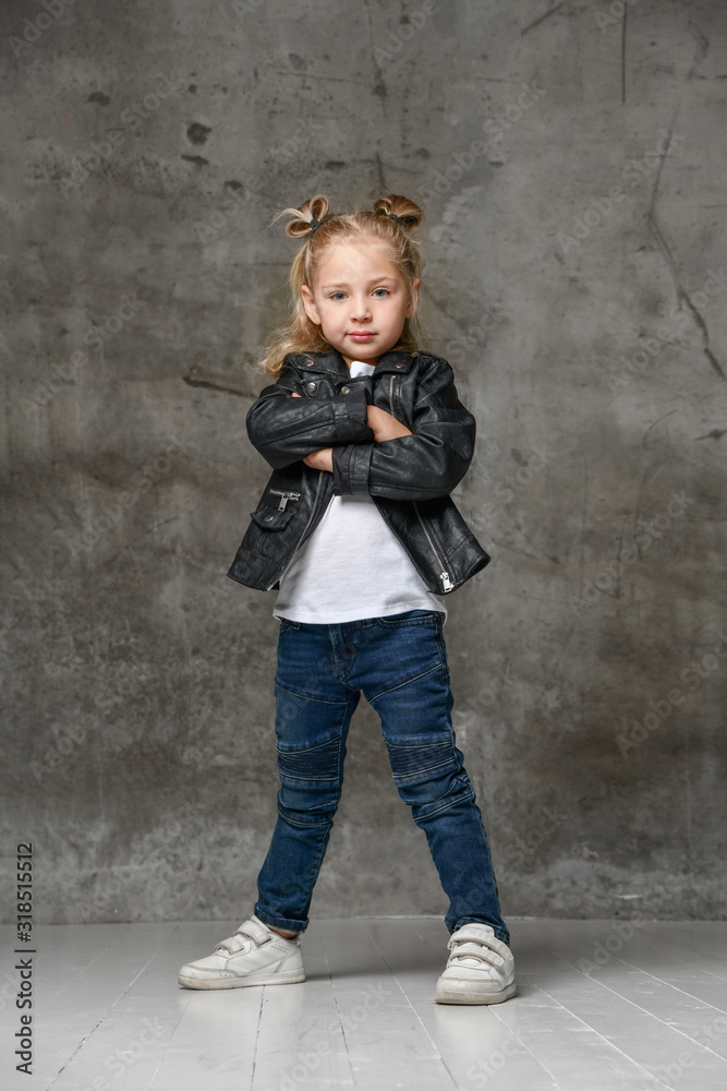 Little cute smiling blond girl in stylish rock style black leather jacket, jeans and white sneakers standing in confident pose Little cute smiling blond girl in stylish rock style black leather jacket, jeans and white sneakers standing in confident pose