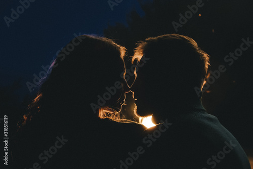 Young couple in love camping tourists sitting by a fire against a tent in the forest with a retro garland, photo with a lot of noise, selective focus