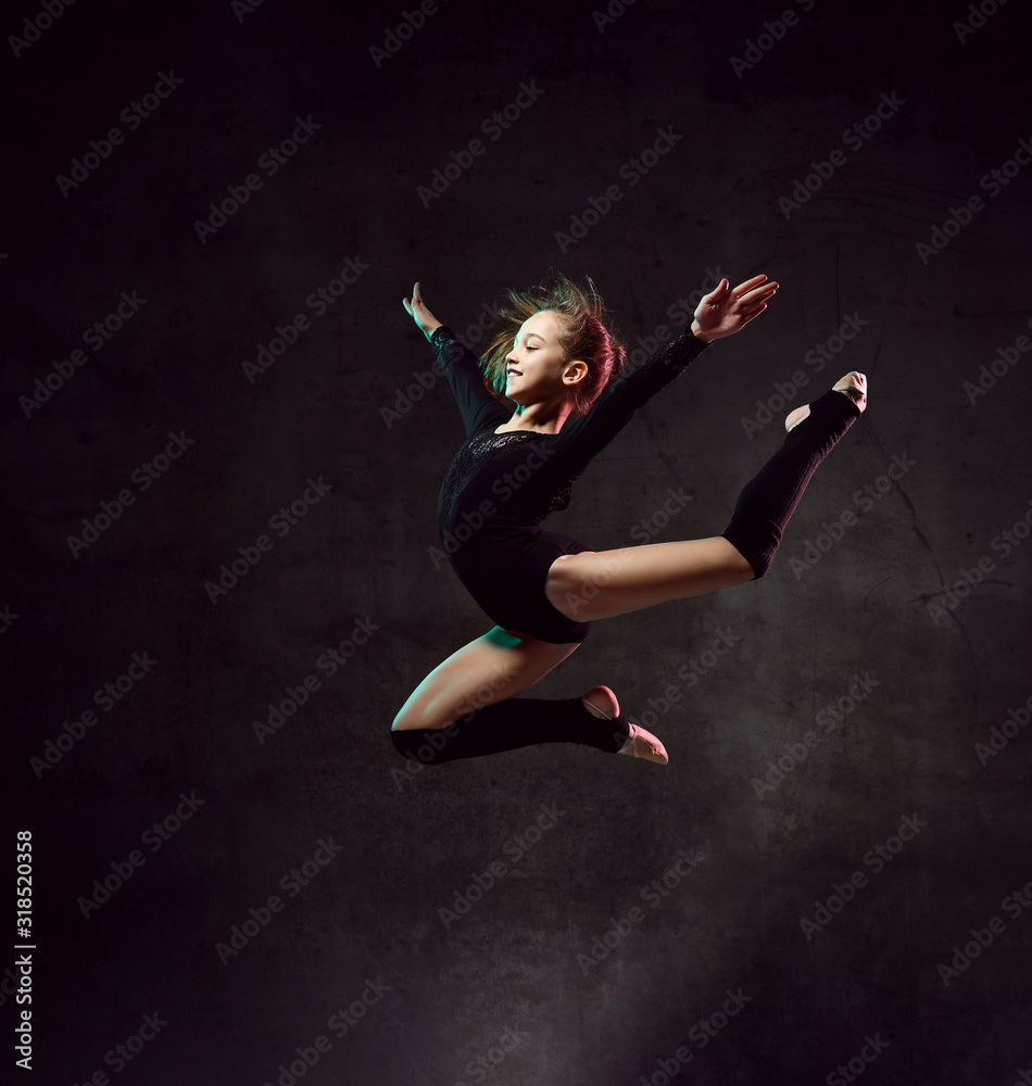 girl gymnast in black sport body and uppers jumping and making dymnastic pose in air over dark background