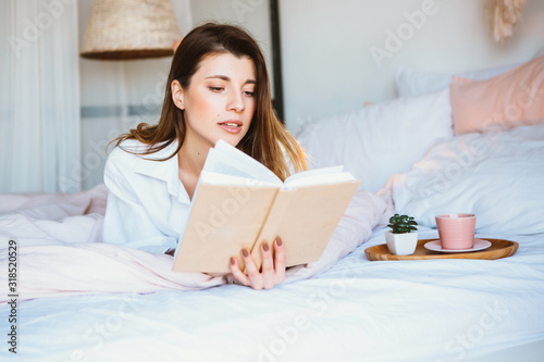 Girl reading a book, drinking coffee or tea, resting in bed.