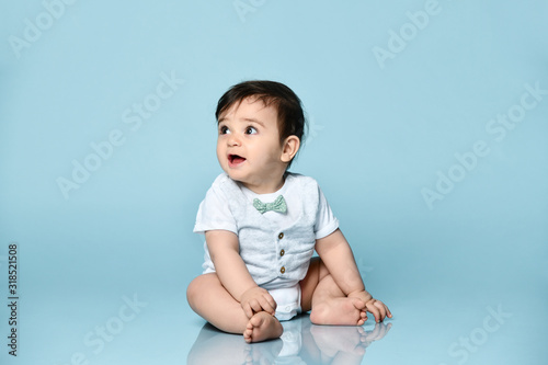 Little toddler in white bodysuit as a vest with bow-tie, barefoot. He is sitting on the floor against blue background. Close up