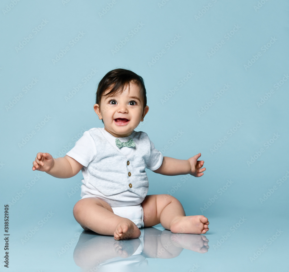 Baby boy in white bodysuit as a vest with bow-tie, barefoot. He smiling, sitting on the floor against blue background. Close up Baby boy in white bodysuit as a vest with bow-tie, barefoot. He smiling, sitting on the floor against blue background. Close up