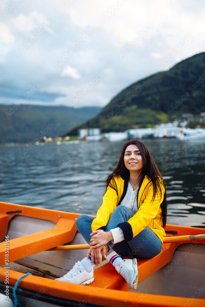 The girl tourist in a yellow jacket  is sitting in a boat against the backdrop of the mountains in the Norway.  Active woman relaxing on the boat by the lake. Travelling, lifestyle, adventure concept.