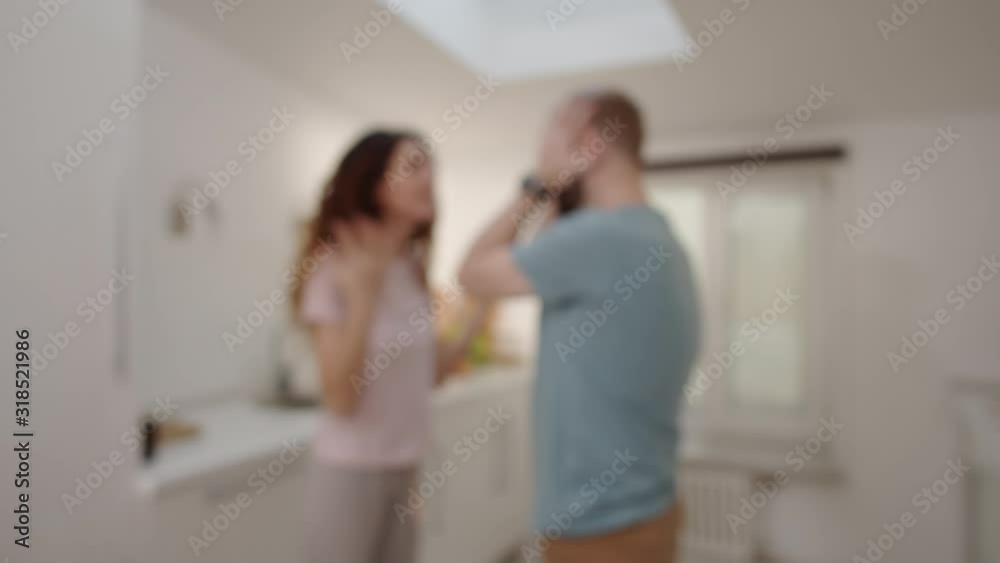 Thigh-up arc shot with blurred effect of husband and wife standing face to face and quarreling at home, nagging woman shouting, reproaching and gesticulating while man is listening and grabbing head Thigh-up arc shot with blurred effect of husband and wife standing face to face and quarreling at home, nagging woman shouting, reproaching and gesticulating while man is listening and grabbing head