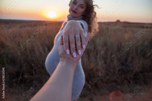 Close-up of the man hand holding the hand with married ring of his pregnant woman against the field and sunset. woman wearing the knitted dress