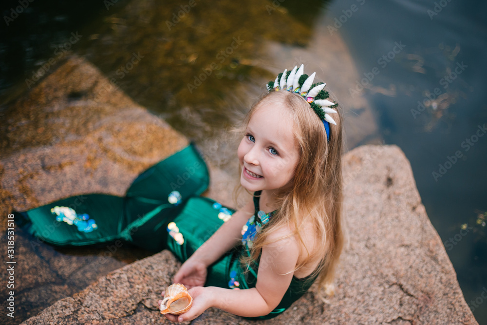 A little girl with white hair with a mermaid tail and shells is sitting on the shore. A little ...