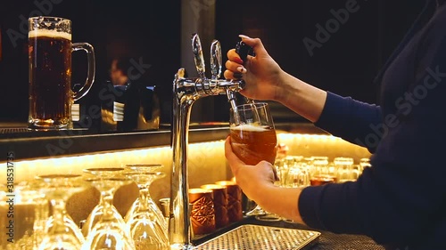 Barmen woman is pouring beer into the clean high glass in stylish bar, close up. Professinonal in blue sweatshirt is opeining the tap by his hand and beer with white foam is flow in the mug. photo