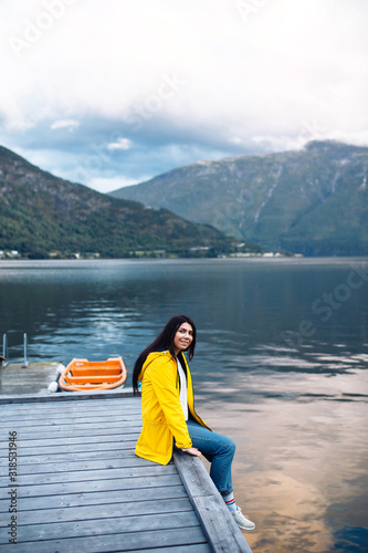 The girl tourist in a yellow jacket posing on the lake in Norway. Active woman relaxing near the lake against the backdrop of the mountains in the Norway. Travelling, lifestyle, adventure, wild nature © maxbelchenko