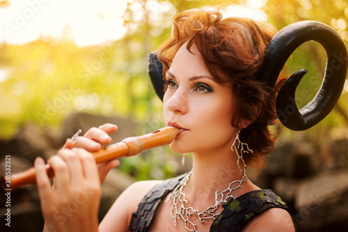 A girl in the image of a faun plays the wooden flute. Model with horns on his head and with furry pants in the woods among the stones photo