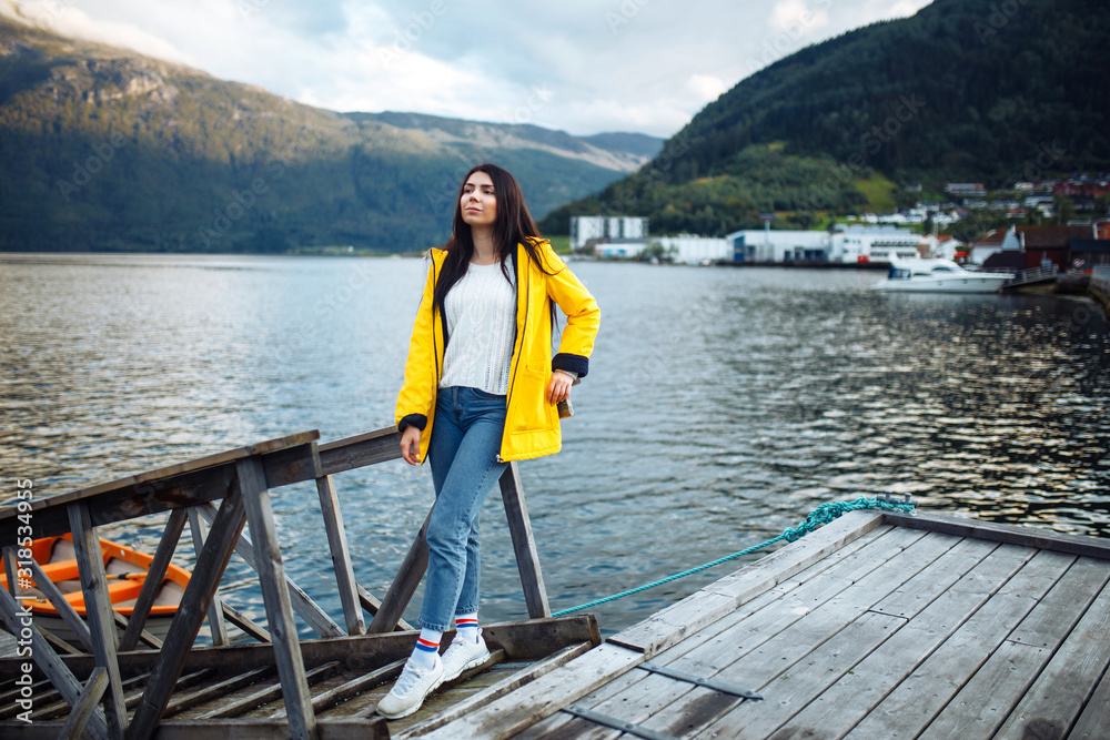 The girl tourist in a yellow jacket posing by the lake in Norway. Active woman relaxing near the boat by the lake against the backdrop of the mountains in the Norway. Travelling, lifestyle, adventure.