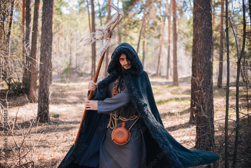 A man with a beard and a mustache in a robe and coat with a staff in his hand. A magician, a shaman conjures in the forest. Fantasy image A man with a beard and a mustache in a robe and coat with a staff in his hand. A magician, a shaman conjures in the forest. Fantasy image