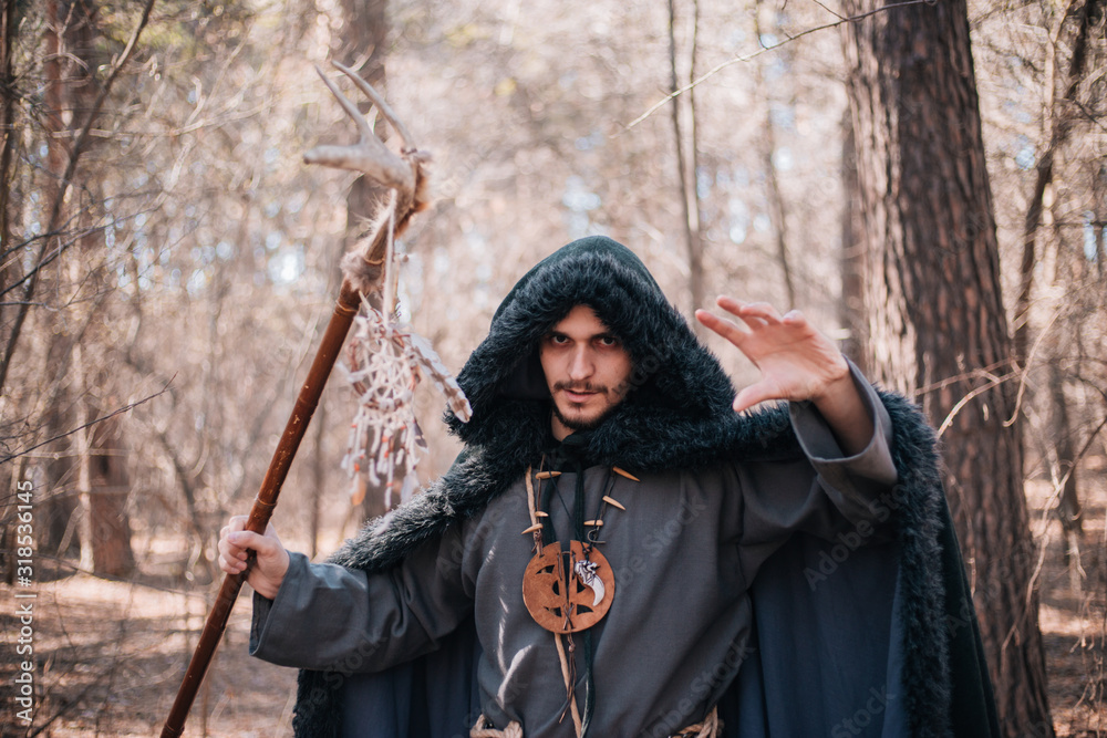 A man with a beard and a mustache in a robe and coat with a staff in his hand. A magician, a shaman conjures in the forest. Fantasy image A man with a beard and a mustache in a robe and coat with a staff in his hand. A magician, a shaman conjures in the forest. Fantasy image