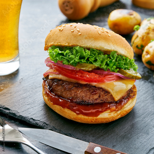 Cheeseburger made of meat burger, cheese, tomato, lettuce, onion and pickled cucumber with baked potato and broccoli served with glass of beer and sauce.