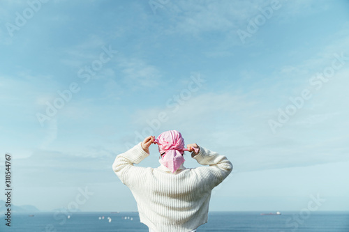 Back view of unknown young woman with cancer lacing pink scarf on her head on the coast.