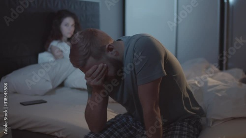 Medium shot of exhausted young Caucasian man sitting on side of bed, with smartphone beside pillow, and rubbing forehead, and angry woman glaring at his back