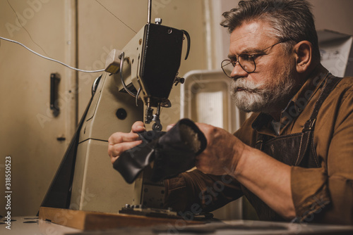 senior, bearded cobbler sewing part of leather shoe on sewing machine