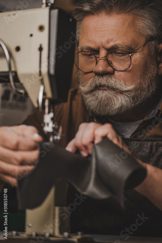 selective focus of attentive, senior shoemaker sewing leather on sewing machine