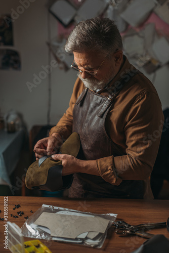 senior cobbler in apron holding piece of genuine leather in workshop