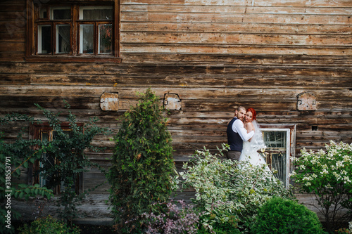 Peaceful secluded newly wed couple hugging in front of an old wooden house wall. Clinging to each other. They are standing between bushes. From afar. photo