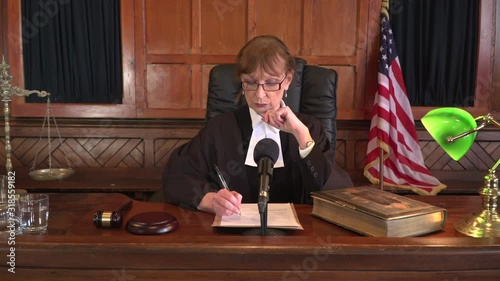 Female Judge sat at the bench in a Court. There is a USA flag behind the magistrate who takes her glasses off