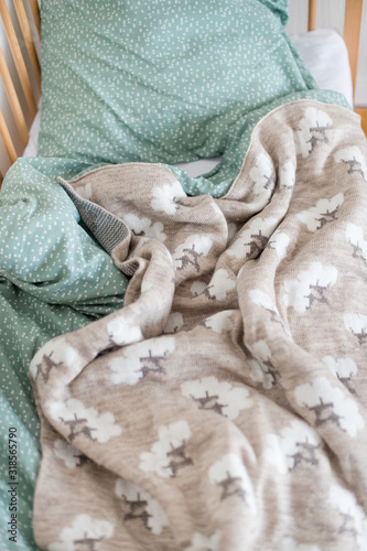 The cot is spread out. We see a crumpled pillow, plaid and blanket. The toy is also in bed. There is a toy baby stroller nearby. photo