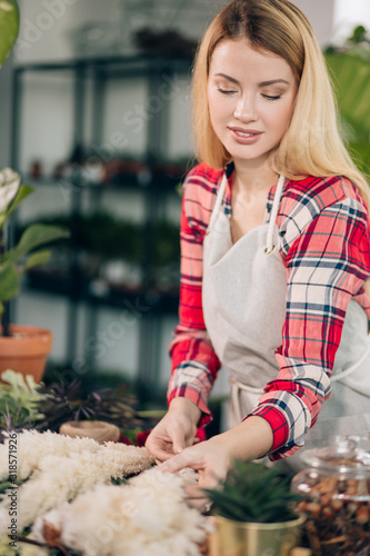 careful florist lady with long blonde hair in red casual checkered shirt and apron stand in her own florist shop, look after plants and flowers