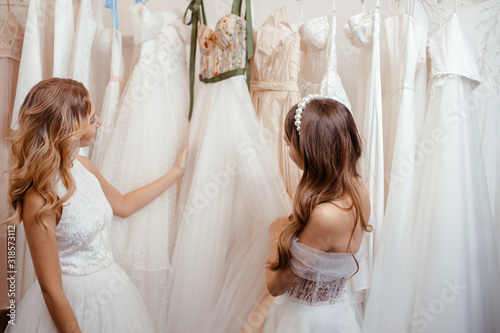 two pretty young women choosing wedding dress in salon, wearing dresses. Interestingly look on dresses, dream about wedding