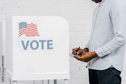 cropped view of african american citizen holding pen and ballot while voting near stand with vote lettering