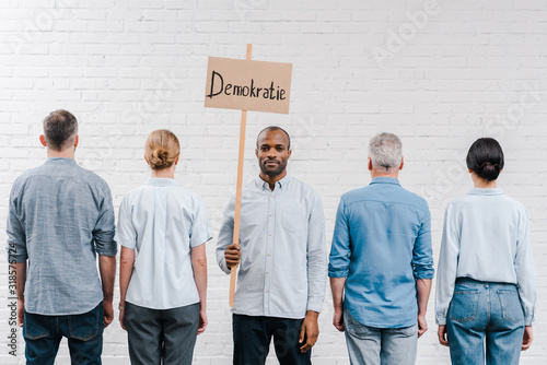 back view of people standing near brick wall near african american man holding placard with demokratie lettering