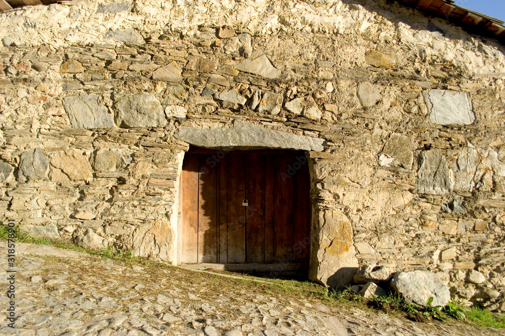 Wooden doors to the ancient building in the georgian village Mestia. A wall of stones of various shapes, from small and flat, to huge rectangular stones. Beam from a single piece of rock.Street tilted