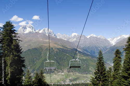 Empty chairs of the cable car on the background of Mount Ushba. The mountain snow peak is partially covered with small clouds against the blue sky. Mountain slopes covered with green forest. Mestia