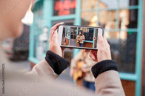 Person Filming Female Musician Busking Playing Acoustic Guitar And Singing To Crowd On Mobile Phone photo